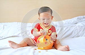 Smiling little Asian baby boy in traditional Chinese dress putting some coins into a piggy bank sitting on bed at home. Kid saving