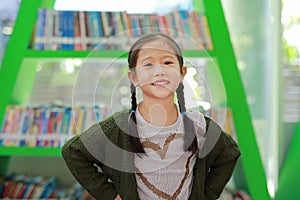 Smiling little Asian child girl against bookshelf at library. Children creativity and imagination concept