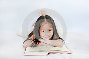 Smiling little Asian child girl with hardcover book lying on bed and looking camera over white background