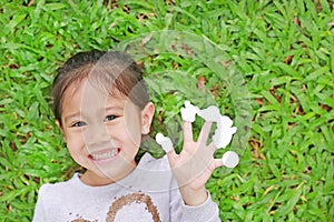 Smiling little Asian child girl lying on green grass lawn with showing empty white stickers on her fingers