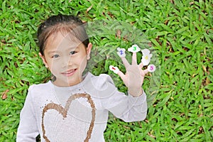 Smiling little Asian child girl lying on green grass lawn with showing white stickers with number one to five on her fingers
