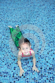 Smiling little Asian child girl in a mermaid suit playing poolside with looking camera