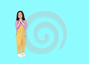 Smiling little Asian child girl in pink-yellow dungarees expression hands implore isolated over cyan background with copy space photo