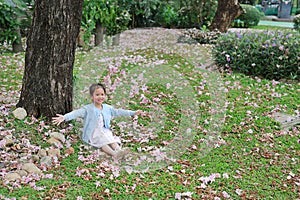 Smiling little Asian child girl sitting on green grass under tree trunk with falling pink flower in the park garden