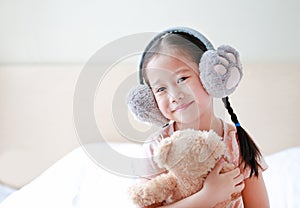 Smiling little Asian child girl wearing winter earmuffs and embracing teddy bear while sitting on the bed at home