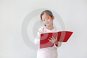 Smiling little Asian child girl writes in a book or notebook with pencil in classroom against white background