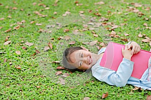 Smiling little Asian girl with book lying on green grass with dried leaves in the summer garden