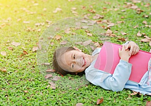 Smiling little Asian girl with book lying on green grass with dried leaves in the summer garden