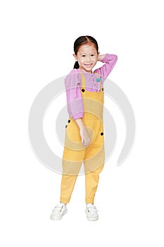 Smiling little Asian kid girl in pink-yellow dungarees poses touched hair keep back isolated on white background. Full length of