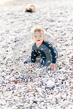 Smiling little baby in overalls sits on a pebble beach. Close-up