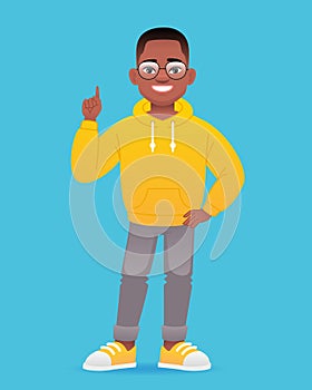 A smiling little black boy stands in glasses and points his finger at something. Cute african school kid in yellow hoodie