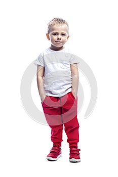 A smiling little blond boy stands with his hands in his pockets. A child in red pants and a white T-shirt. Isolated on white