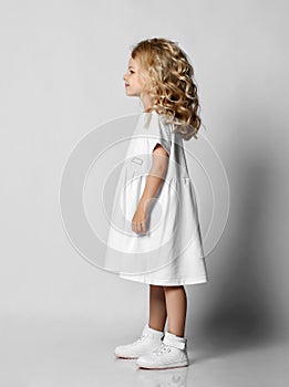 Smiling little blonde curly kid girl preschooler in white casual dress and sneakers is standing side to us