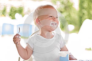 Smiling little blonde toddler boy eating delicious yogurt sitting outdoor