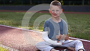 smiling little boy with broken hand is sitting outdoors on sports ground on summer day. 9 years child with a fractured