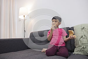 Smiling little boy eating bread and watch TV on comfortable sofa in the living room. Enjoying playing game on digital smart