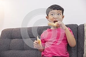 Smiling little boy eating bread and watch TV on comfortable sofa in the living room. Enjoying playing game on digital smart