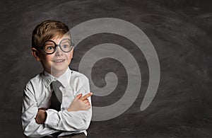 Smiling Little Boy in Glasses over School Black Board. Funny Happy Child with crossed Arms pointing Finger to Copy Space