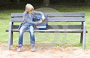 Smiling little boy opening his backpack. Child sitting on a wooden bench. Outdoor. Education, school concept