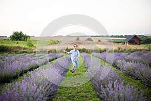 Smiling little boy running on the lavender field. Funny child in glasses dressed in a blue shirt and a straw hat running on a gras