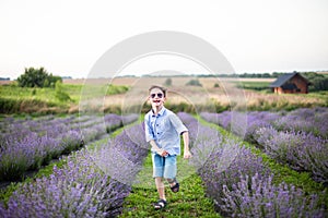 Smiling little boy running on the lavender field. Funny child in sunglasses dressed in a blue shirt and a straw hat running on a g