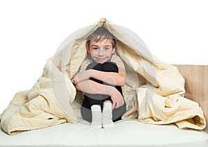 Smiling little boy sitting on a bed under a blanket and hugging her knees