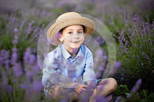Smiling little boy sitting on the lavender field. Funny child dressed in a blue shirt and a straw hat sitting between lavender flo