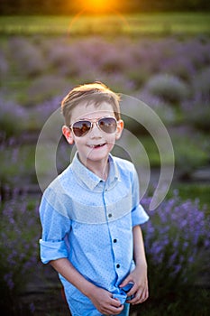 Smiling little boy standing on the lavender field. Funny child in sunglasses dressed in a blue shirt and a straw hat standing betw