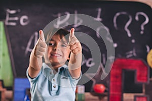 Smiling little boy with thumbs up in the classroom against to blackboard background