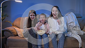 Smiling little boy with two teenage girls watching late night TV show and eating popcorn from big bowl