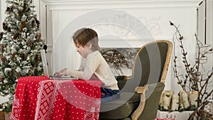 Smiling little boy typing letter to Santa Claus on a laptop near the Christmas tree