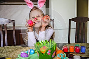 Smiling little brunette girl with bunny ears and painted nose sitting at home at the table holding painted Easter eggs