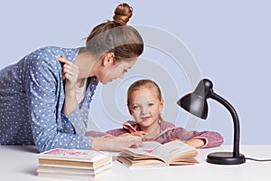 Smiling little charming girl sits at table and her mother help her to do homework task, try to learnn poem together, uses reading