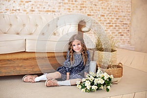 Smiling little curly hair girl with a bouquet of jasmine flowers sits by the sofa in cozy  autumn living room. Scandinavian interi