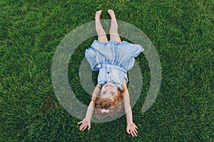 Smiling little cute child baby girl in denim dress lie on green grass lawn, raise hands up in park. Mother, little kid