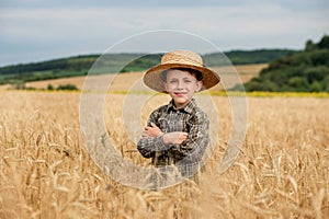 A smiling little farmer boy in a plaid shirt and straw hat poses for a photo in a wheat field. Heir of farmers