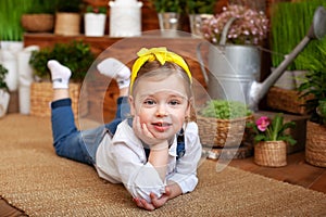 Smiling little girl with bandage her hair lying on wooden floor around green houseplants and flowers. Concept of childhood. Garden