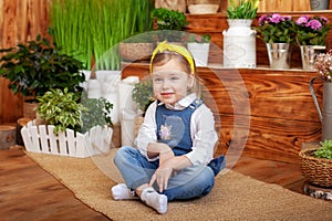 Smiling little girl with bandage her hair sits on wooden floor around green houseplants and flowers. Concept of childhood. Gardeni