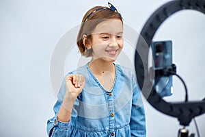 Smiling little girl blogger in casual denim dress use selfie led lamp and smartphone
