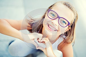 Smiling little girl in with braces and glasses showing heart with hands