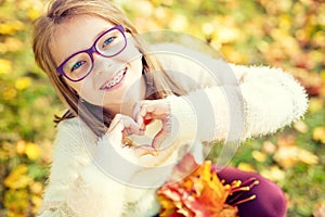 Smiling little girl with braces and glasses showing heart with hands.Autum time