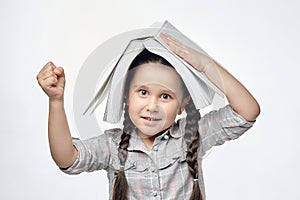 Smiling little girl with braided pigtails shows her fist while holding a large book over her head