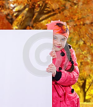 Smiling little girl child in autumn clothes jacket coat and hat holding a blank billboard banner white board.
