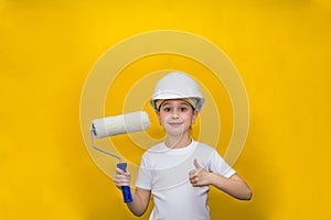 Smiling little girl in a construction white helmet holds a paint roller on a yellow background. concept of renovation