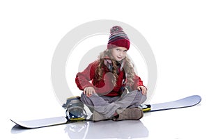 Smiling little girl with curly hairstyle wearing knitted sweater, scarf, hat and gloves sitting on blue snowboard, isolated