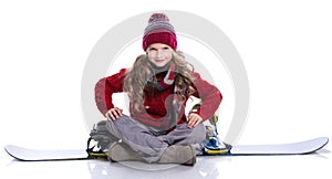 Smiling little girl with curly hairstyle wearing knitted sweater, scarf, hat sitting on blue snowboard, isolated on white.