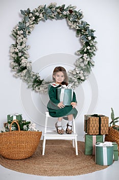 Smiling little girl in dress sit on chair with gifts in decorated living room for Christmas. Beautiful little girl with big fir Xm