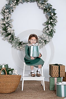 Smiling little girl in dress sit on chair with gifts in decorated living room for Christmas. Beautiful little girl with big fir Xm