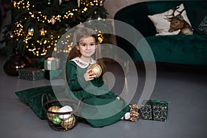 Smiling little girl in dress sit on floor with gift and Xmas balls toy by Christmas tree with glowing garland lights. Beautiful sm