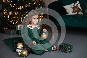 Smiling little girl in dress sit on floor with gift and Xmas balls toy by Christmas tree with glowing garland lights. Beautiful sm